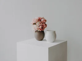 Vases Manufacture Un vase marron avec des fleurs roses et un vase blanc en céramique sont posés sur un cube blanc sur un fond clair et sobre.