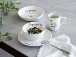 La coupe Statement « Lieblingsmensch » [La personne la plus chère à mon cœur] avec des myrtilles, l’assiette assortie ainsi qu’un mug à anse et une cuillère sur une table.