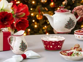 Théière et chauffe-théière de Toys Delight. Une table festive avec une théière, un chauffe-théière, une tasse, des céramiques de Noël, des biscuits et un sapin décoré en arrière-plan.