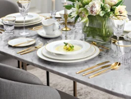 Place setting with grand royal, metrochic and chateau septfontaines Elegant table set on a marble surface with gold-rimmed plates, gold flatware, crystal glasses, a vase of white flowers, and a soup served on the central plate.