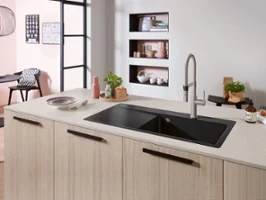 Un évier de cuisine Subway Style dans un îlot de cuisine clair avec une robinetterie Une cuisine moderne avec un évier noir sur un îlot de cuisine en bois clair, un robinet, une plante et divers objets.
