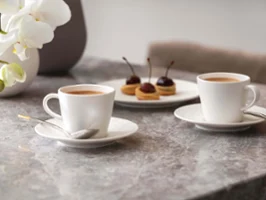 Une tasse à expresso Manufacture Rock Blanc avec de la pâtisserie Deux tasses à expresso Manufacture et une assiette de pâtisseries sur une table en marbre avec des orchidées en arrière-plan.