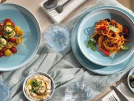 Une table dressée avec des pâtes, de la salade, du houmous, des olives et des couverts sur des assiettes bleues de la collection Perlemor Aqua  et une nappe claire.