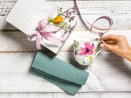 Une main attrape une tasse Mariefleur de Villeroy & Boch à côté d'une boîte cadeau. Une main attrape une tasse Mariefleur de Villeroy & Boch à côté d'une boîte cadeau avec un ruban rose et des fleurs, posée sur une surface en bois avec un tissu bleu-vert.