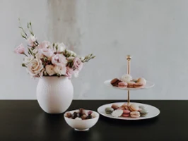 Vase Manufacture et présentoir MetroChic Un vase blanc avec des fleurs roses, un plateau à plusieurs étages avec des desserts et une coupe de chocolats sur une table sombre.