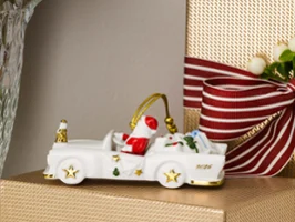 Une petite voiture de Noël en céramique chargée de cadeaux trône sur un paquet cadeau doré, à côté d'un vase rempli de fleurs rouges et blanches.