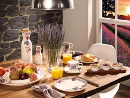 Un couvert de petit-déjeuner avec la vaisselle Villeroy & Boch Artesano Provençal Lavande, orné de fruits, d'un croissant, de céréales et de jus d'orange. Un vase de lavande trône élégamment au centre de la table. La fenêtre offre une vue pittoresque d'un coucher de soleil sur un champ de fleurs violettes.