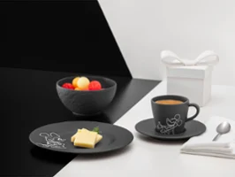 Vaisselle noire de la collection Manufacture Mickey à côté d'un coffret cadeau blanc Vaisselle noire de la collection Manufacture Mickey avec fruits, tartine beurrée et café, à côté d'un coffret cadeau blanc, d'une serviette et d'une cuillère sur un fond divisé.