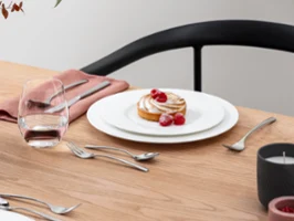 Royal combiné avec des délices sucrés Un dessert garni de framboises est posé sur une assiette Royal avec des couverts sur une table en bois. À côté se trouvent un verre d'eau, une serviette rose et une composition florale.