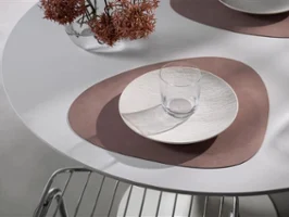 Lifestyle Accessoires avec Manufacture Un verre et une assiette sont posés sur un set de table abstrait marron de Villeroy & Boch Lifestyle Accessoires sur une table blanche, avec en arrière-plan une chaise en métal et un vase avec des fleurs séchées.
