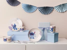 Un set de napperons avec des assiettes, des tasses et des bols sur le thème de "Stitch" Un set de napperons avec des assiettes, des tasses et des bols sur le thème de "Stitch" sur des boîtes bleues, avec une boîte cadeau rayée et une décoration.