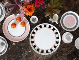 Memphis dans toutes ses facettes Service de table Memphis de Villeroy & Boch avec des assiettes décoratives, une tasse, des tasses et des prunes sur une surface en marbre brun décorée de fleurs brillantes.