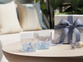 Deux verres transparents Like Aqua et un coffret cadeau blanc Deux verres transparents Like Aqua et un coffret cadeau blanc avec un ruban bleu sur une table ronde ; canapé et coussins en arrière-plan.
