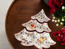 Vue de dessus d'un set de 3 coupelles à snacks en porcelaine en forme de sapin de Noël de la collection Toy's Delight sur une table en bois foncé, contenant des biscuits en forme d'étoiles et de cœur, à côté d'un cadeau rouge avec un ruban rayé vert et blanc et des fleurs rouges