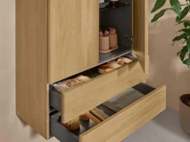 Un placard haut Antao en structure bois dans une salle de bains Une armoire de salle de bains Villeroy & Boch en bois clair avec deux portes et deux tiroirs ouverts adossés à un mur naturel.
