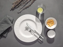 Couverts Piemont sur une assiette Une assiette blanche avec une fourchette et un couteau, un verre d'eau avec du citron, un bol de crackers et une serviette de table sur une table grise.