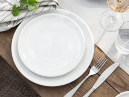 Un Artesano- set de table de Villeroy & Boch avec des assiettes blanches, des couverts, une serviette rayée avec un motif de fleurs, un verre de rosé, du pain sur une planche et un bol avec des artichauts.
