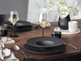 Une table à manger moderne en bois avec un service de table Manufacture Une table à manger moderne en bois avec un service de table Manufacture, des verres à vin, des couverts et un vase avec des fleurs blanches.