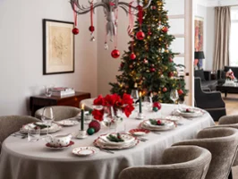 De la vaisselle Toy’s Delight dans une salle à manger décorée pour Noël Une salle à manger décorée pour Noël avec une table dressée pour huit personnes. La table est décorée avec de la vaisselle festive, des bougies et des fleurs rouges. À côté, il y a un sapin de Noël avec des ornements.