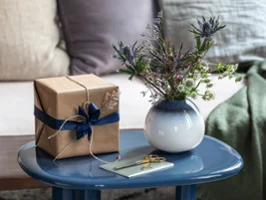 Sur une table bleue, un vase de lave blanc Sur une table bleue se trouvent un cadeau enveloppé dans du papier brun avec un ruban bleu, un vase Lave blanc avec des fleurs et une carte.