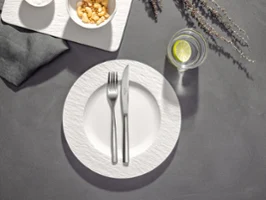 Une assiette avec une fourchette croisée et un couteau dessus, à côté d'un verre d'eau avec du citron vert et deux bols de snacks sur une table.
