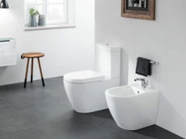 Une salle de bains moderne avec des toilettes blanches et un bidet sur un sol carrelé. Sur le côté gauche, il y a un tabouret en bois et une petite armoire, et sur le mur, une fenêtre et une œuvre d’art encadrée.