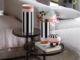Deux vases à rayures noires et blanches et un récipient rond sont posés sur des tables en verre à côté d'un canapé gris, l'un des vases contient des fleurs