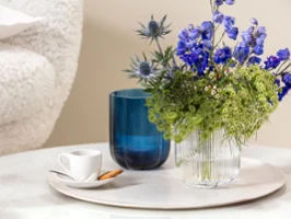 Une petite tasse For me blanche avec un biscuit sur une soucoupe, à côté d'un vase Fleur bleu et d'un vase Fleur transparent Une petite tasse For me blanche avec un biscuit sur une soucoupe, à côté d'un vase Fleur bleu et d'un vase Fleur transparent avec des fleurs bleues et vertes sur une surface de marbre ronde.