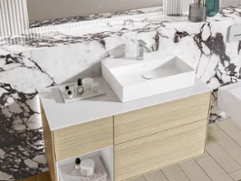Un plan de toilette en bois clair avec une vasque angulaire devant un revêtement en marbre avec un grand miroir.