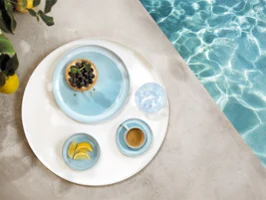 Un plateau rond avec de la vaisselle de la collection Perlemor Aqua et un verre d'eau, au bord d'une piscine.