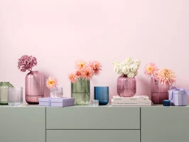 Une série de vases de la collection Fleur, de formes et de tailles variées, disposés sur une étagère. Une série de vases aux couleurs pastel de la collection Fleur, de formes et de tailles variées, avec des fleurs roses, blanches et couleur pêche, posés sur une étagère avec des livres empilés et des petites boîtes.