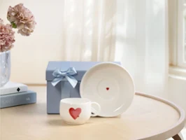 Tasse With Love avec soucoupe comme idée cadeau Tasse blanche With Love avec motif cœur rouge, soucoupe assortie et boîte cadeau bleue avec nœud sur une table en bois près de la fenêtre.