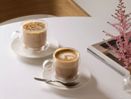 Deux mugs en verre Artesano de Villeroy & Boch sur une table Deux tasses en verre Artesano de Villeroy & Boch sur une table : l’une remplie de café glacé, l’autre de latte, avec une cuillère, une pile de magazines et une fleur rose dans un vase.