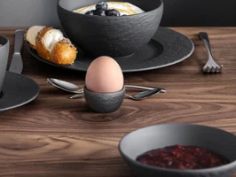 Manufacture Couvert de petit-déjeuner sur une table en bois Un ensemble de petit-déjeuner avec du café, du jus, un croissant, de la confiture et un œuf mollet sur une table en bois, des fleurs vertes sont visibles.