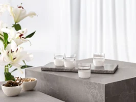Quatre verres à eau Manufacture blancs sur une table en béton, accompagnés de lys blancs dans un vase et des coupes Manufacture contenant des snacks.