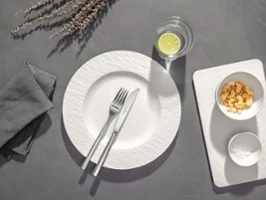 Une assiette blanche avec une fourchette et un couteau, un verre d'eau avec du citron et un plateau avec des céréales et un bol sur un fond gris.