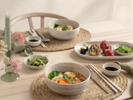 Une table dressée pour deux personnes avec des bols de ramen, des rouleaux de sushi, des edamame, des baguettes et des sauces à tremper de la collection Perlemor sur des sets de table tissés.