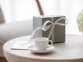 Une tasse à expresso et une sous-tasse Afina avec un emballage cadeau Une tasse Afina blanche sur une sous-tasse, une boîte cadeau avec un ruban et un bouquet de fleurs blanches sur une table ronde.