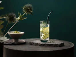 Un verre La Divina de Villeroy & Boch servi avec style Un verre La Divina avec des tranches de concombre et du romarin, sur une surface en pierre avec un fond vert. Une paille en verre Artesano est plantée dans la boisson et une coupe d’olives est posée à côté.