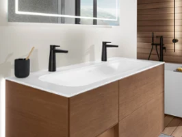Deux lavabos Finion avec des robinetteries noires Un lavabo moderne avec un plan de toilette blanc, deux robinets noirs, un porte-brosse à dents noir avec une brosse à dents et des placards en bois en dessous. Un grand miroir avec éclairage intégré est accroché au mur au-dessus.