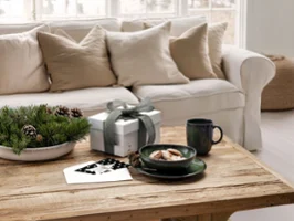 Une table basse en bois avec une tasse Lave green, des snacks, des plantes et du papier devant un canapé blanc avec des coussins beiges.