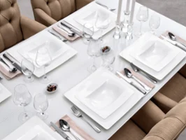 Une table de salle à manger dressée avec de la vaisselle Modern Grace Une table de salle à manger joliment décorée avec de la vaisselle blanche carrée de la série Modern Grace de Villeroy & Boch, des couverts argentés, des verres à vin et des serviettes beiges. Des chaises beiges rembourrées entourent la table. Trois bougeoirs sont placés au centre.
