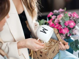 Mug Letters avec la lettre M dans la main d’une femme Une femme prend en main une tasse Letters avec un « M ». À côté, une autre femme est assise avec un smartphone.