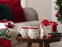 Tasses de Noël festives avec coffret cadeau et décorations de Noël Deux tasses de Noël, un coffret cadeau et des décorations de Noël sur une petite table à côté d'un salon décoré pour les fêtes.