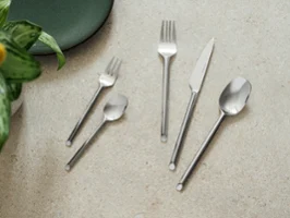 Cinq couverts like de like.by Villeroy & Boch- fourchette, couteau, cuillère, fourchette à dessert et cuillère à café Cinq couverts like. Like. by Villeroy & Boch - fourchette, couteau, cuillère, fourchette à dessert et cuillère à café - sont disposés sur une surface en pierre claire à côté d'une assiette verte et d'une plante.