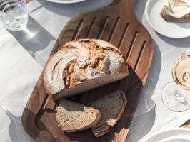 Une miche de pain avec deux tranches repose sur une planche en bois Artesano de Villeroy & Boch. Sur la table, il y a des assiettes, des serviettes, un verre d'eau, un bol d'olives et un verre de vin.