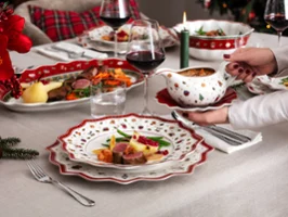 Une table de Noël festive dressée avec de la vaisselle Toy’s Delight Une table festive est dressée avec de la vaisselle de Noël Toy’s Delight de Villeroy & Boch et des assiettes de viande, de légumes et de sauce sont présentées. Une personne tient une saucière, des verres à vin rouge et une bougie allumée sont également présents.