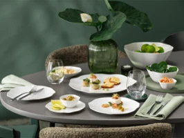 Une table ronde dressée avec la vaisselle blanche de la collection Pura, comprenant des assiettes à entrée, des verres, des serviettes et un centre de table composé de plantes vertes.