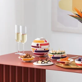 La Boule Miami with Manufacture Blanc Collier Vase in modern arrangement Table moderne avec La Boule Miami, assortiment de snacks, deux coupes de champagne et un vase blanc Manufacture Collier avec des fleurs orange.