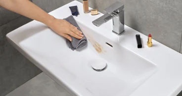 Une main nettoie un lavabo dans la salle de bains avec un chiffon gris et enlève une tache de maquillage. Différents produits de maquillage sont posés sur le bord du lavabo.